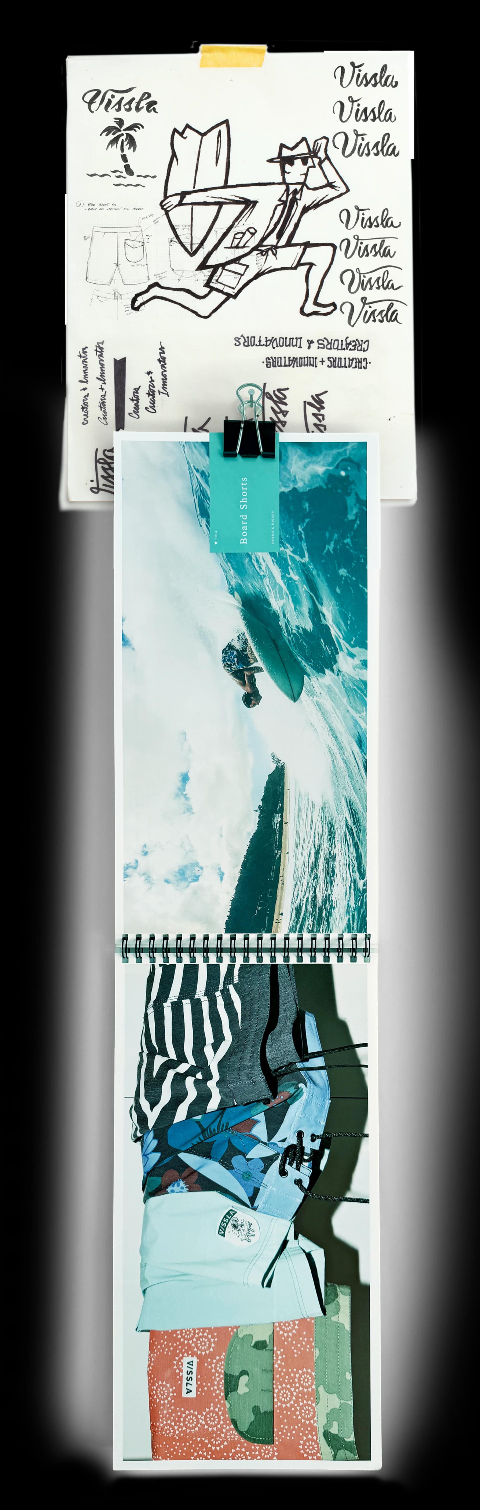 Inked logo studies and a drawing of a running figure carrying a surfboard, clipped above a spiral-bound boardshort lookbook open to a surf photograph and a stack of shorts.