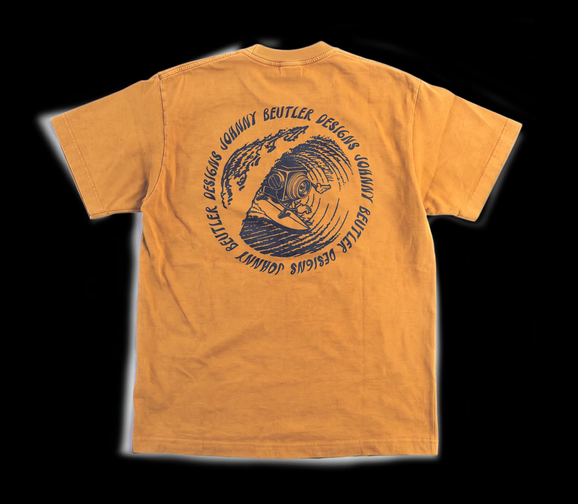 A mustard t-shirt printed on the back with a circular Johnny Beutler Designs mark: a camera-headed figure riding a wave inside a ring of hand-drawn type.