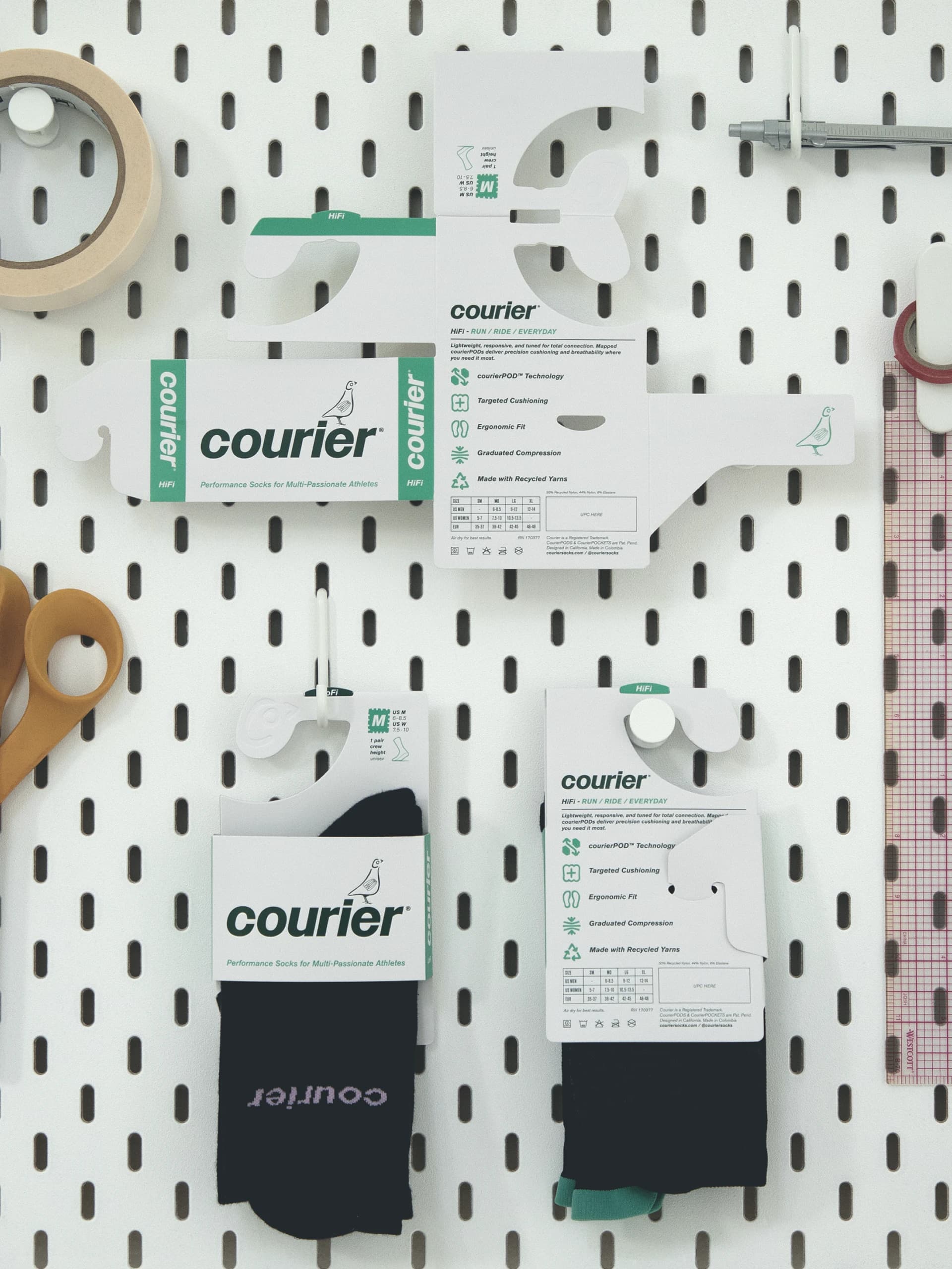 Courier packaging on a white pegboard: a die-cut hanger card unfolded flat above two black socks in their packs.