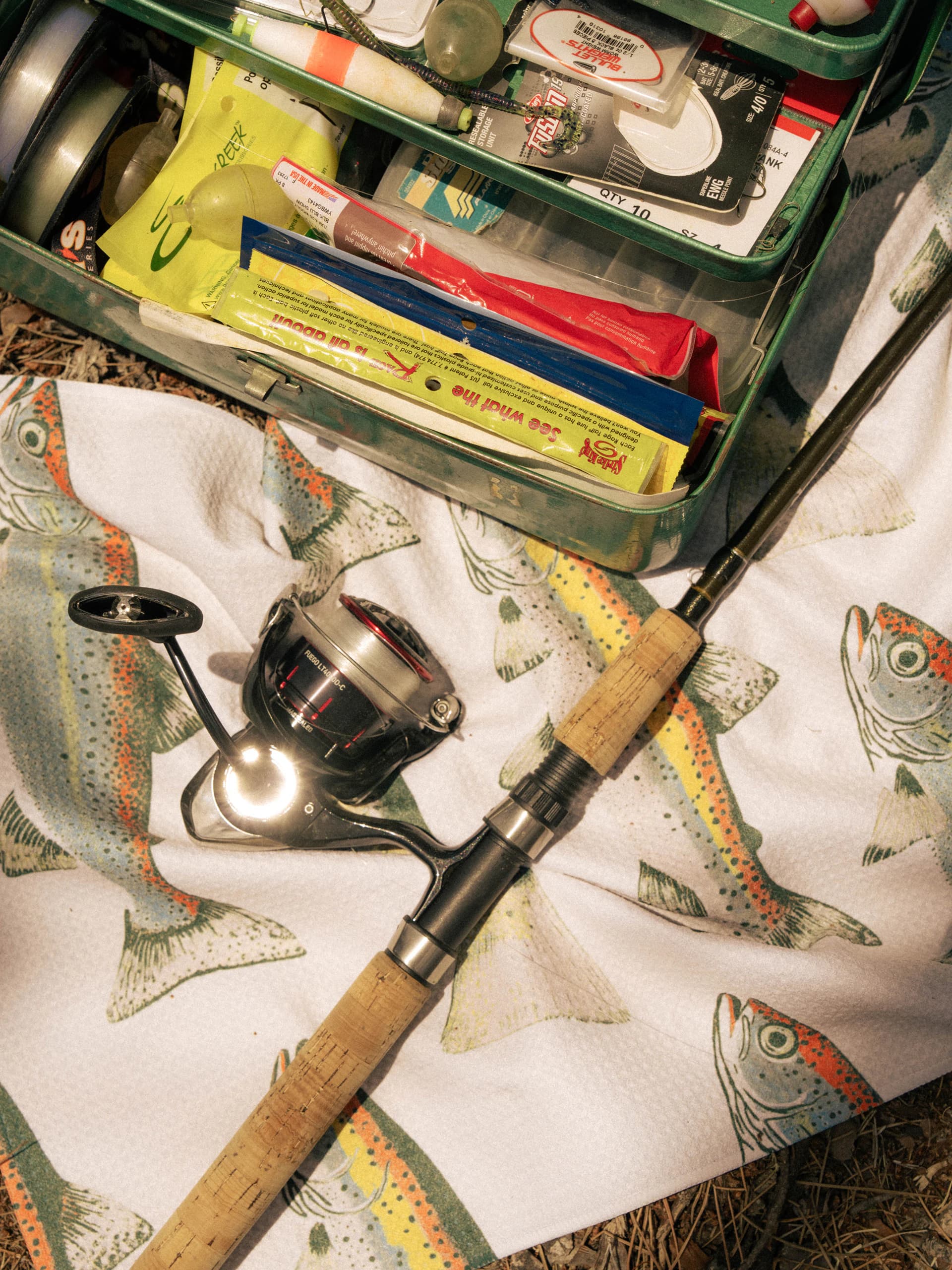 A spinning rod and reel on a towel printed with rainbow trout, beside an open tackle box.