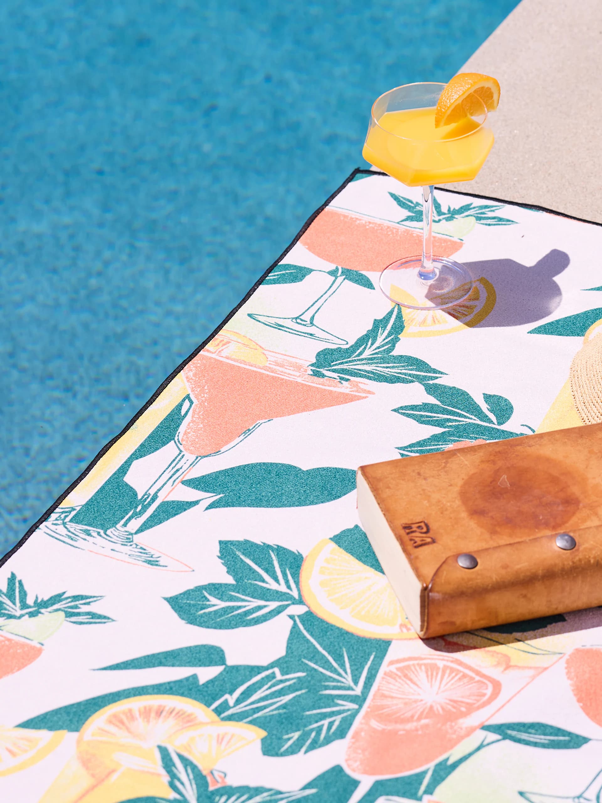 A poolside towel printed with pink cocktails, citrus slices and palm leaves, a glass of orange juice and a leather case resting on it.