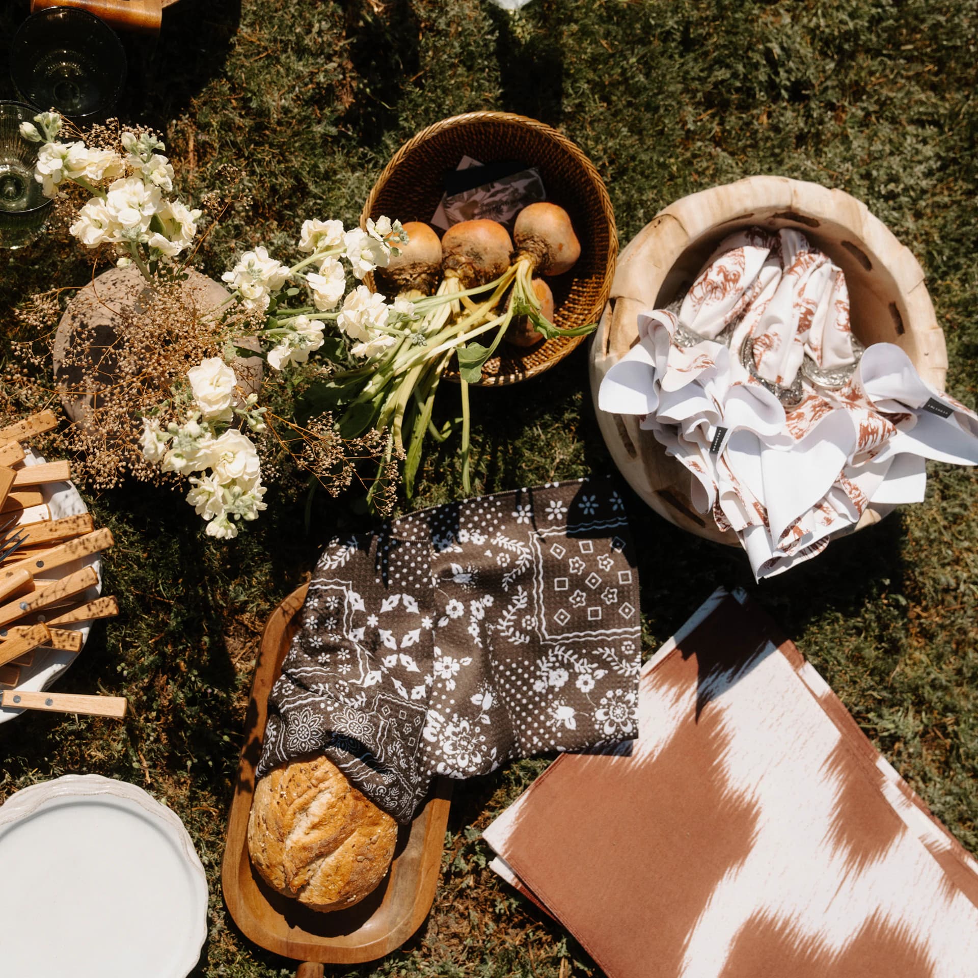A picnic spread on the grass from above: bread on a brown bandana-print towel, napkins in a bowl and a brown cowhide-print towel.