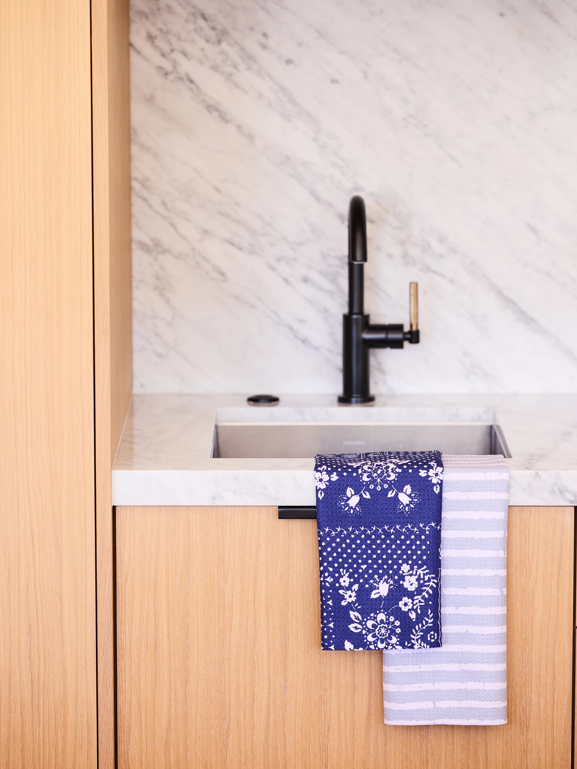 Two towels hanging from a kitchen sink under a marble backsplash, one a navy floral and one a pale blue stripe.