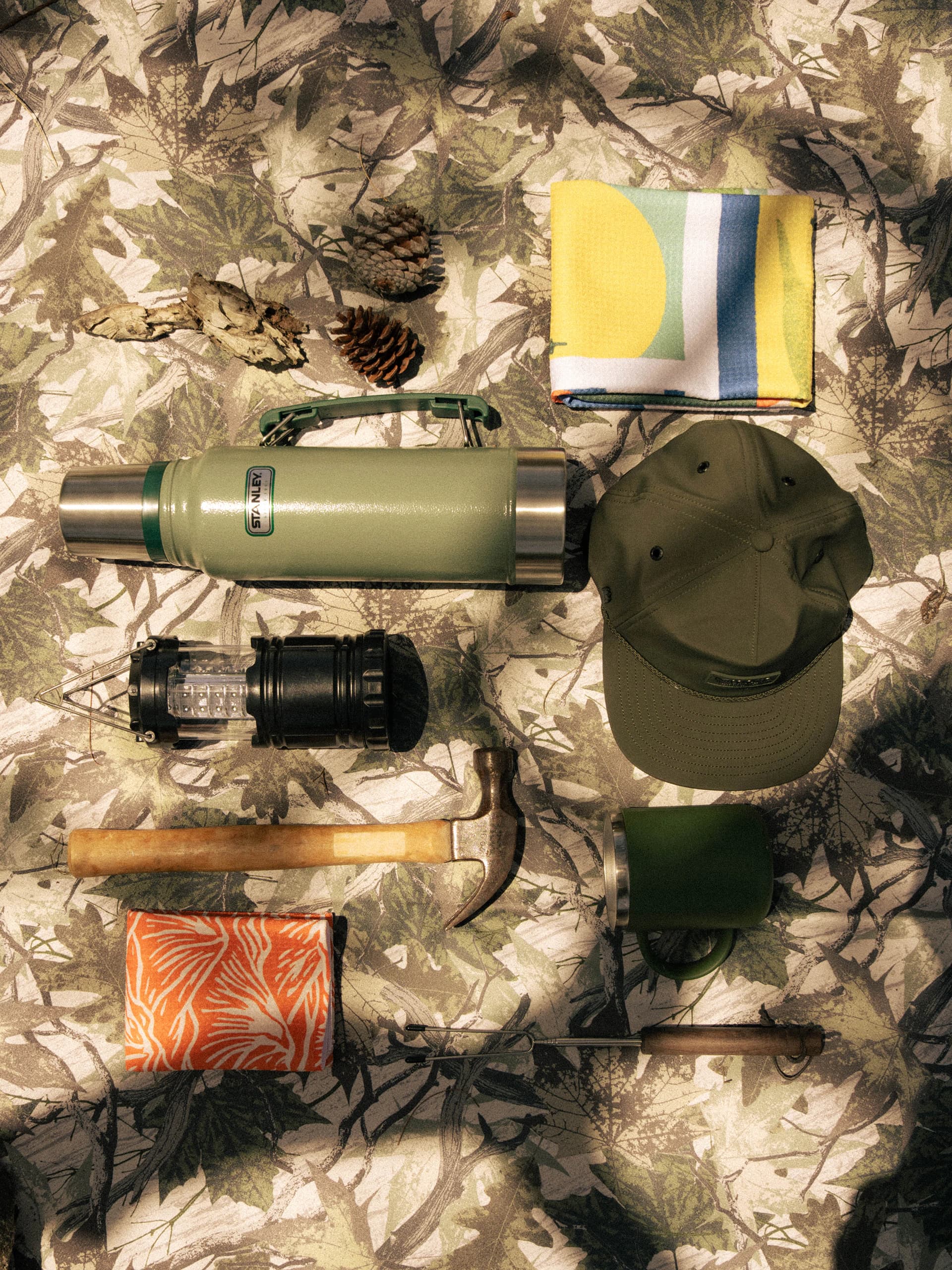 A camping flat lay on a camouflage-print towel: a Stanley flask, cap, lantern, hammer, mug and two folded printed towels.