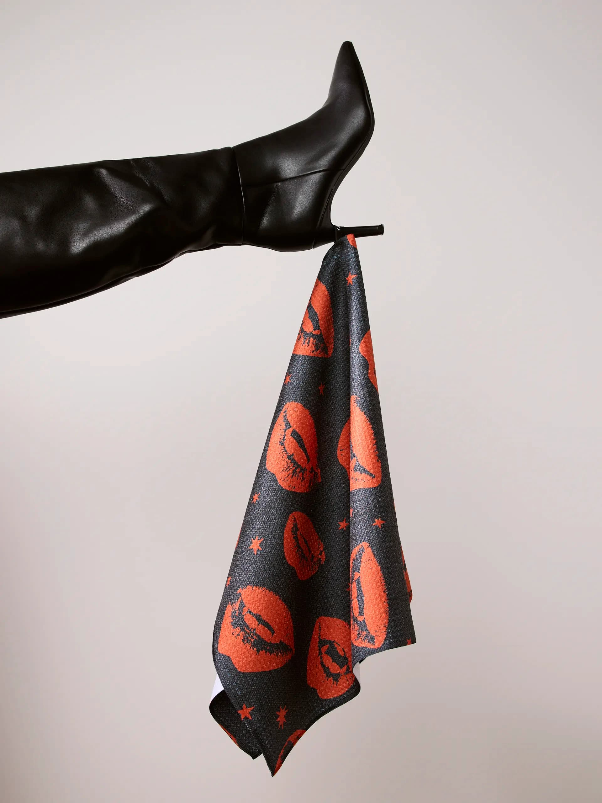 A towel printed with red lips and small stars on charcoal, hanging from the heel of a black stiletto boot.