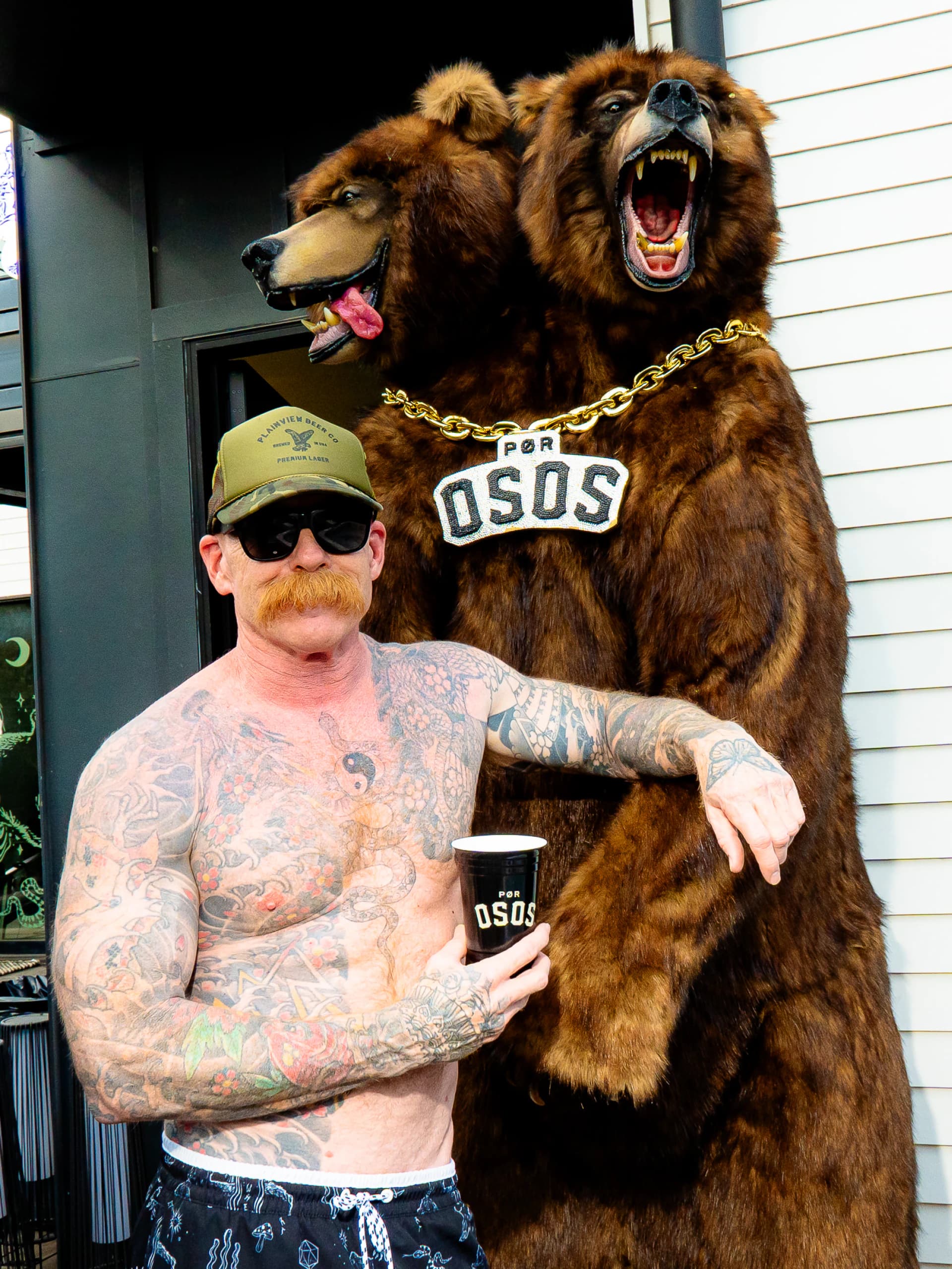 A tattooed man in a cap and sunglasses posing with a stuffed two-headed bear wearing a gold Por Osos chain.