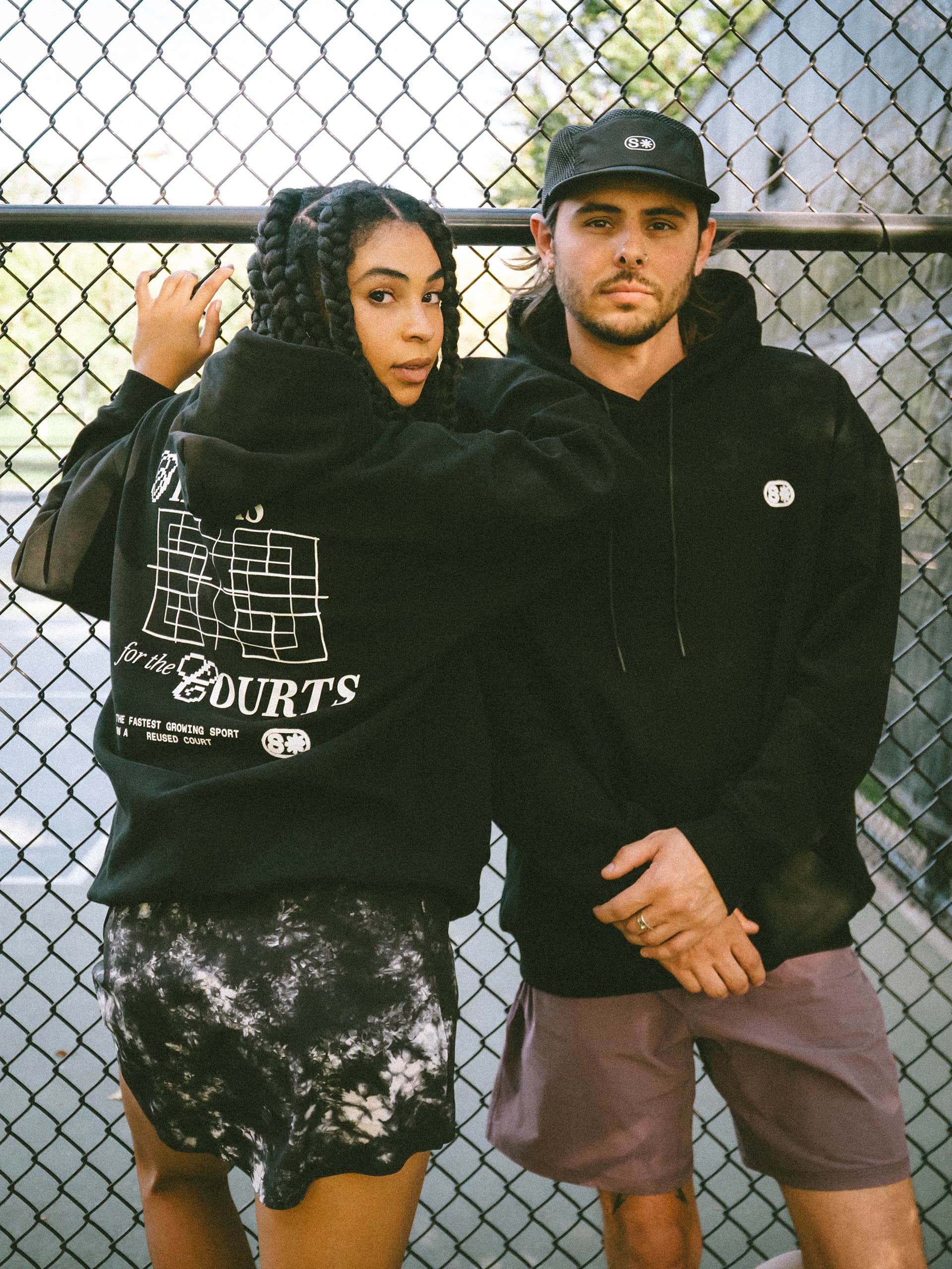 A woman in braids and a man in a Stack cap lean on a chain-link fence in black hoodies, hers showing the Thanks for the Courts back print.
