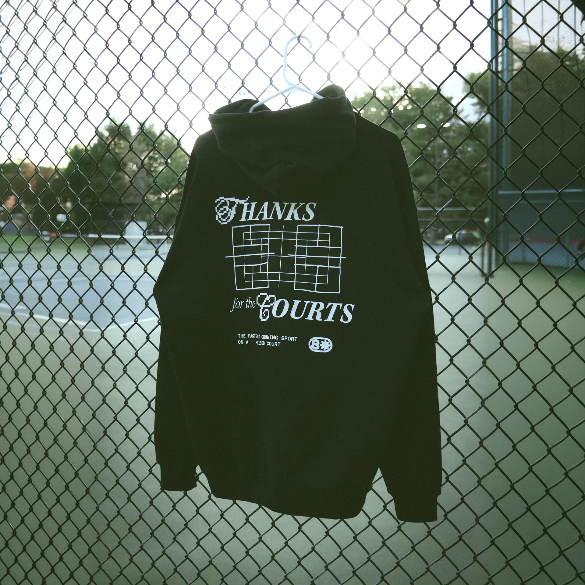 A black hoodie on a hanger against a court fence, its back printed in white: Thanks for the Courts, over a hand-drawn court grid.