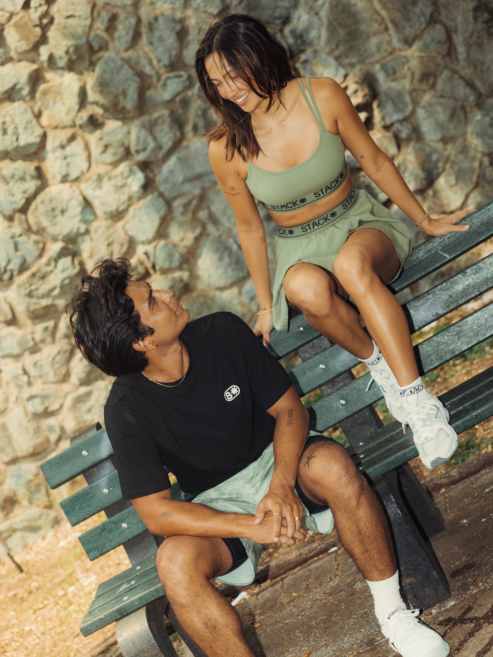 A man in a black tee sits on a green park bench while a woman in a sage Stack set perches on its back, smiling down at him.