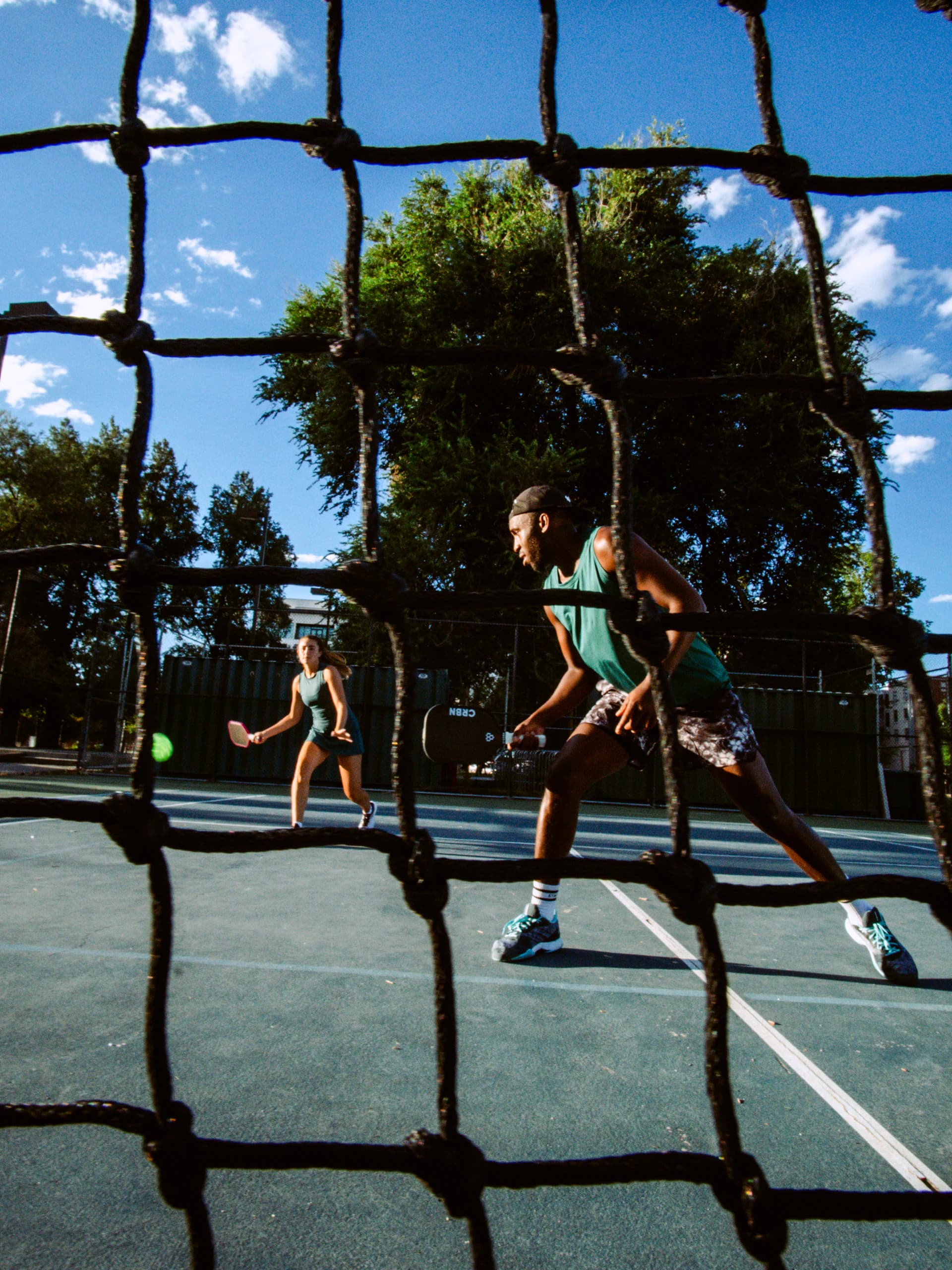 Seen through a court net, a man in a green tank and tie-dye shorts lunges for a shot while his partner waits at the far side.