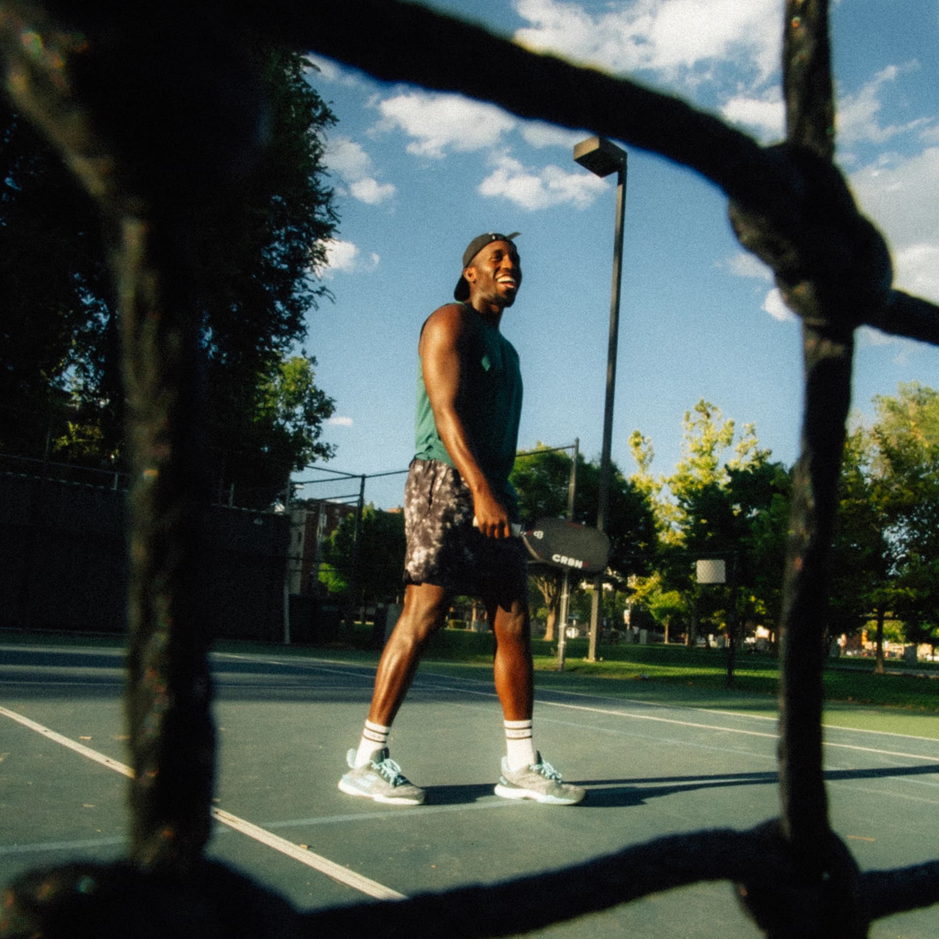 The same player, laughing, walks the court with his paddle, framed through the dark knots of a net.